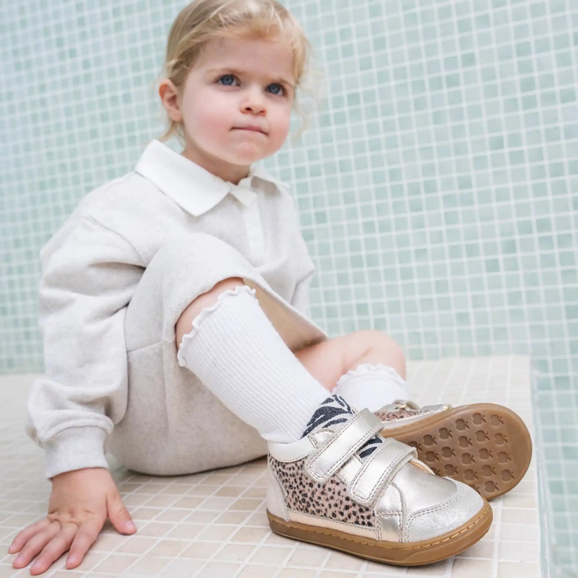 Petite fille en chemise col crème et chaussettes hautes blanches, portant des baskets en argent et à motif léopard avec deux velcros.