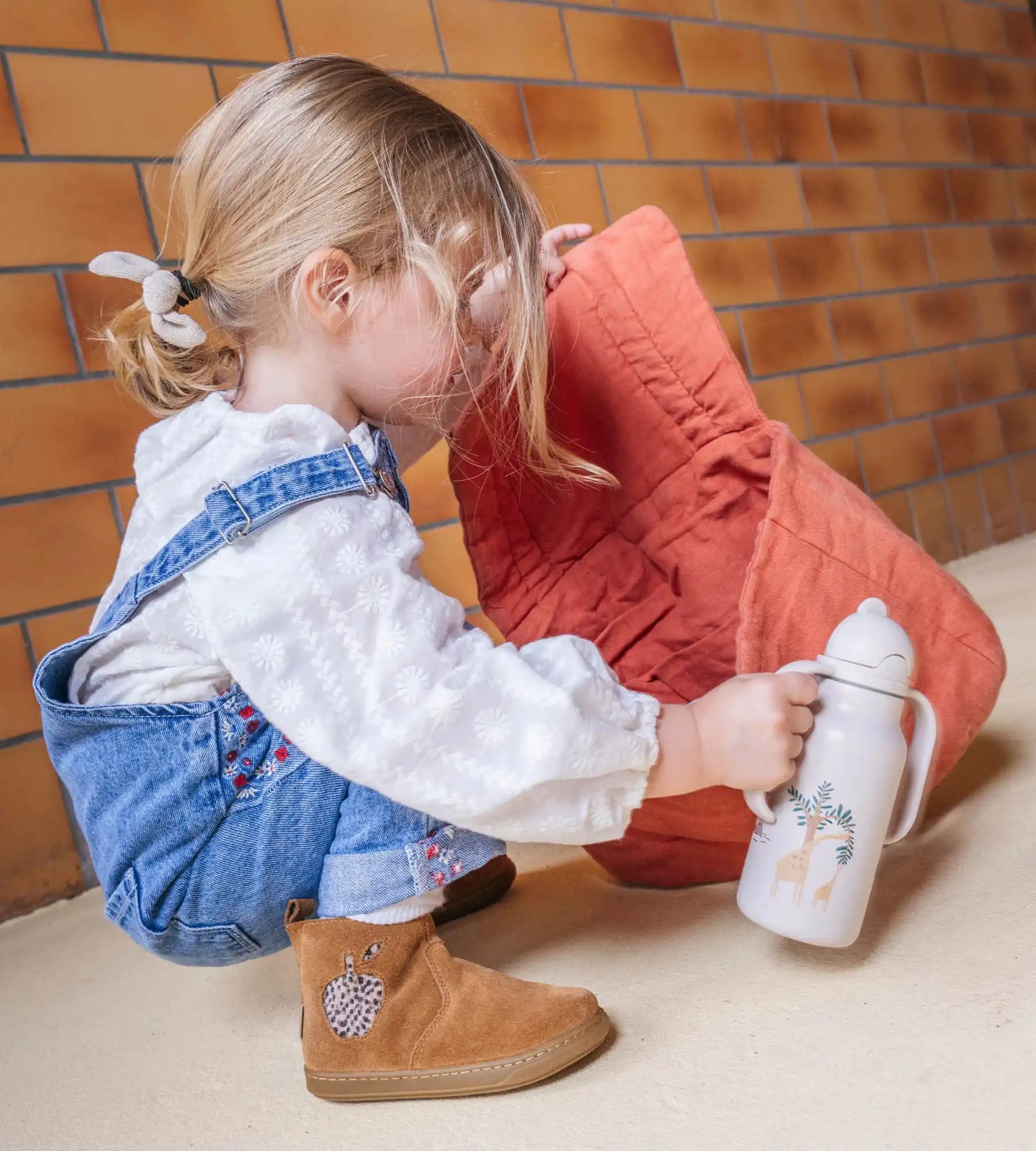 Tout-petit en salopette en jean et bottes marron tenant une tasse à bec verseur.