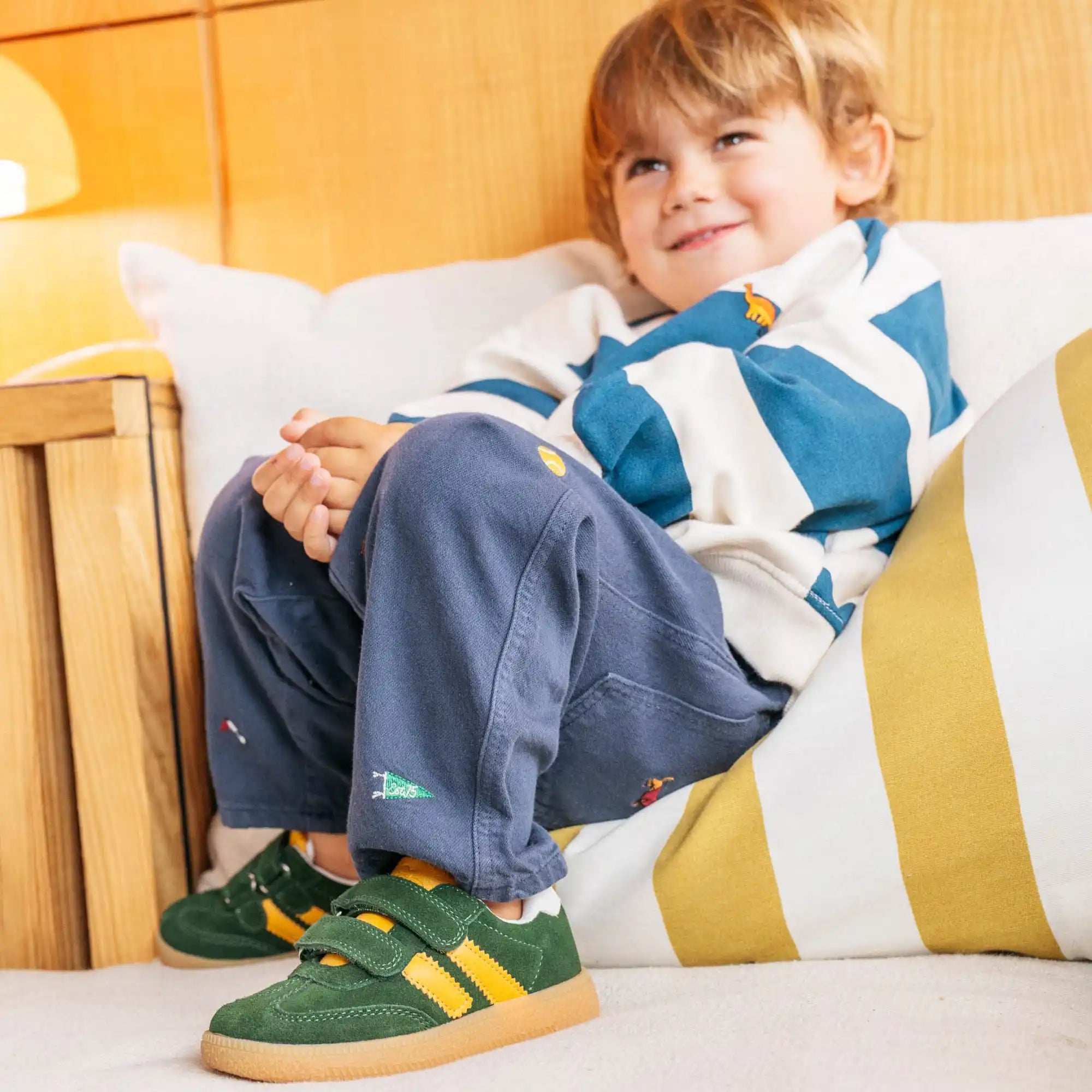Un jeune garçon avec les cheveux blonds, portant une chemise à manches longues rayée bleue et blanche, un pantalon bleu avec des détails brodés colorés, et des baskets vertes avec des rayures jaunes et des bandes velcro.